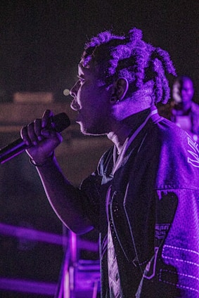 A close-up of a person speaking softly into a sleek microphone, bathed in deep blue and amber light.