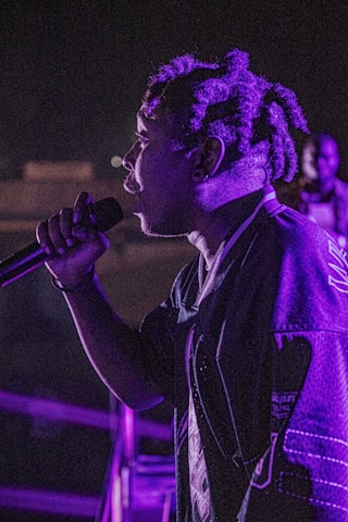 A person holding a microphone stands in profile under dramatic lighting, with purple hues illuminating the scene. The background is dark, highlighting the individual's presence and attire.