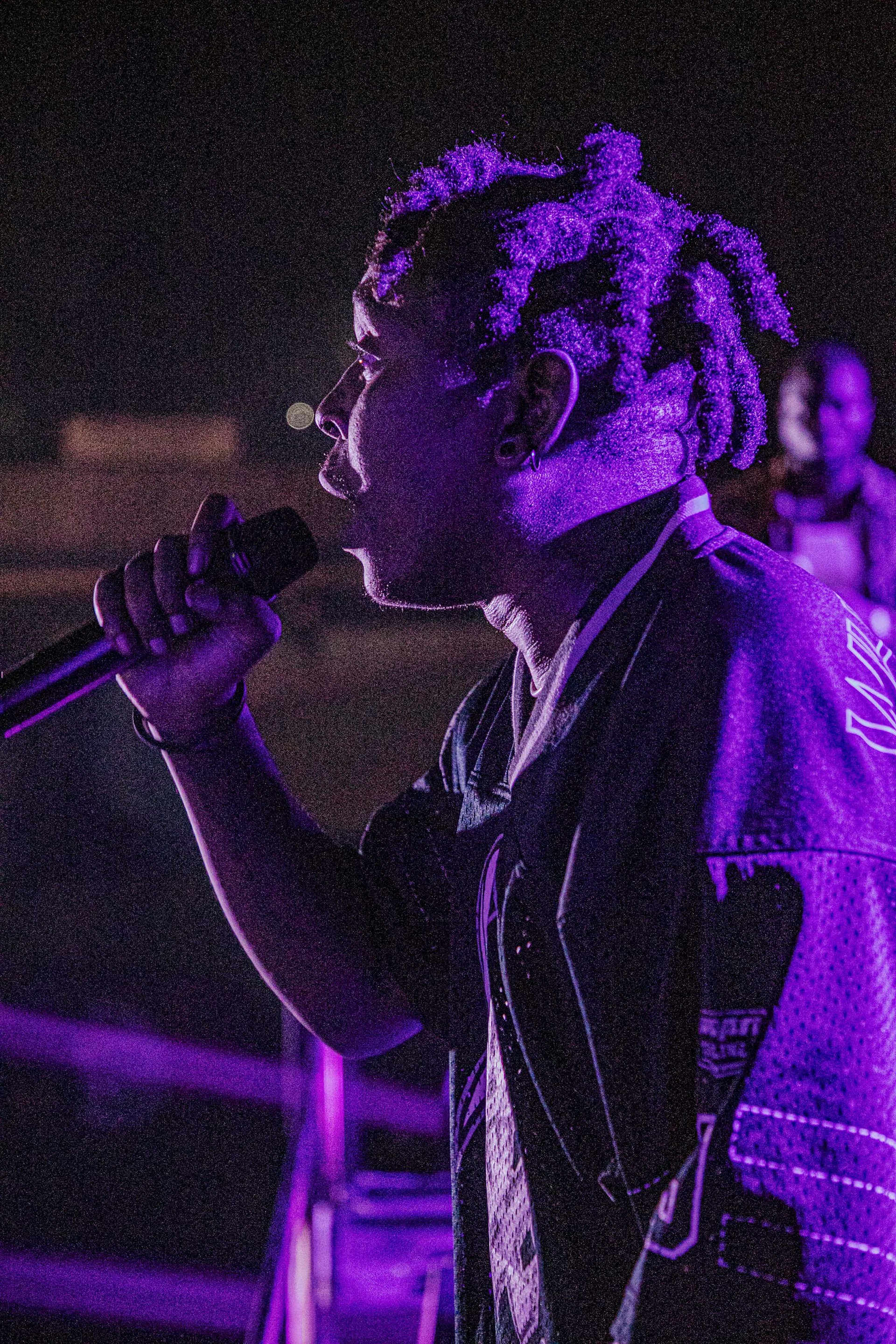 A person holding a microphone stands in profile under dramatic lighting, with purple hues illuminating the scene. The background is dark, highlighting the individual's presence and attire.