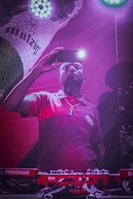 A person in a red shirt is holding a smartphone with its flashlight on, capturing a moment or taking a selfie. They stand in front of DJ equipment, illuminated by purple and pink lighting. A colorful, branded backdrop is partially visible in the background.