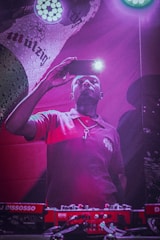 A person in a red shirt is holding a smartphone with its flashlight on, capturing a moment or taking a selfie. They stand in front of DJ equipment, illuminated by purple and pink lighting. A colorful, branded backdrop is partially visible in the background.