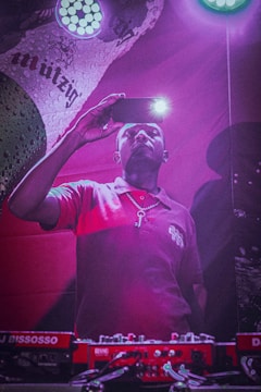 A person in a red shirt is holding a smartphone with its flashlight on, capturing a moment or taking a selfie. They stand in front of DJ equipment, illuminated by purple and pink lighting. A colorful, branded backdrop is partially visible in the background.