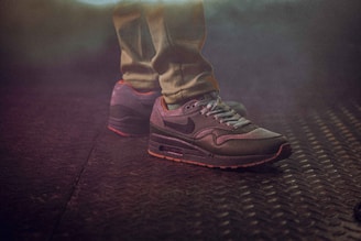 A pair of athletic shoes worn on feet, standing on a textured metal surface with a foggy or smoky ambiance. The lighting is dim and moody, highlighting the design of the sneakers.