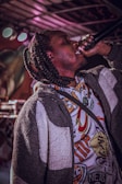 A person with braided hair passionately singing or speaking into a microphone while wearing a colorful, graphic-patterned shirt and a textured jacket. The background features colorful lights and indistinct forms, suggesting a stage or concert environment.
