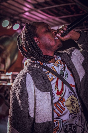 A person with braided hair passionately singing or speaking into a microphone while wearing a colorful, graphic-patterned shirt and a textured jacket. The background features colorful lights and indistinct forms, suggesting a stage or concert environment.