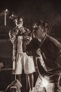 A musician passionately performs on stage, holding a microphone close to their mouth. Behind the performer, a person is focused on capturing the moment with a smartphone, wearing casual attire. The setting appears to be a live concert, with dim lighting adding to the atmospheric feel.