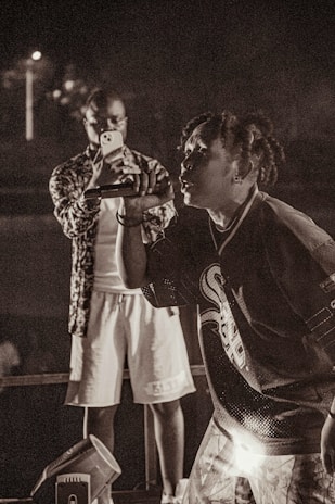 A musician passionately performs on stage, holding a microphone close to their mouth. Behind the performer, a person is focused on capturing the moment with a smartphone, wearing casual attire. The setting appears to be a live concert, with dim lighting adding to the atmospheric feel.