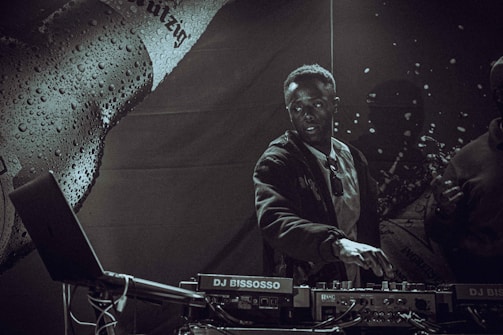 A DJ is performing on stage, interacting with music equipment. The atmosphere is dark and moody, with a focus on the DJ who appears concentrated and engaged. The background is decorated with a textured surface, possibly part of the stage setup or promotional material. Lighting is low, creating a dramatic effect with contrasting shadows.
