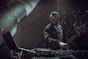 A DJ is performing on stage, interacting with music equipment. The atmosphere is dark and moody, with a focus on the DJ who appears concentrated and engaged. The background is decorated with a textured surface, possibly part of the stage setup or promotional material. Lighting is low, creating a dramatic effect with contrasting shadows.