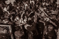 A large group of people gathered closely together, engaging energetically with raised arms and expressions of excitement. The scene appears to be a lively crowd or celebration.