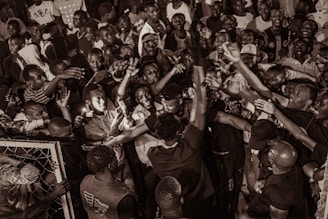 A large group of people gathered closely together, engaging energetically with raised arms and expressions of excitement. The scene appears to be a lively crowd or celebration.