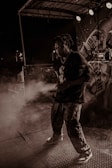 A person is performing on stage under dramatic lighting with a backdrop featuring various designs. There is a haze of smoke or fog surrounding them, creating an atmospheric effect. The setting appears to be during a night event with spotlights adding to the ambiance.