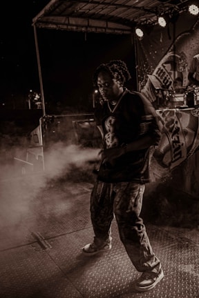 A person is performing on stage under dramatic lighting with a backdrop featuring various designs. There is a haze of smoke or fog surrounding them, creating an atmospheric effect. The setting appears to be during a night event with spotlights adding to the ambiance.