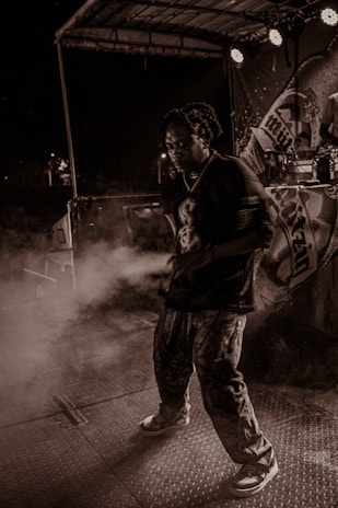 A person is performing on stage under dramatic lighting with a backdrop featuring various designs. There is a haze of smoke or fog surrounding them, creating an atmospheric effect. The setting appears to be during a night event with spotlights adding to the ambiance.