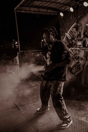 A person is performing on stage under dramatic lighting with a backdrop featuring various designs. There is a haze of smoke or fog surrounding them, creating an atmospheric effect. The setting appears to be during a night event with spotlights adding to the ambiance.