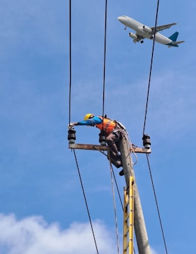 A worker in a safety helmet and orange vest is climbing a utility pole, adjusting cables or equipment. An airplane is flying in the clear blue sky above, close to the utility lines.