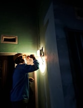 A person wearing a hard hat is inspecting a light fixture on a wall using a flashlight. The setting is dimly lit, suggesting an indoor environment, possibly under maintenance or construction. Shadows cast by the flashlight create a dramatic effect.