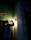 A person wearing a hard hat is inspecting a light fixture on a wall using a flashlight. The setting is dimly lit, suggesting an indoor environment, possibly under maintenance or construction. Shadows cast by the flashlight create a dramatic effect.