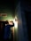 A person wearing a hard hat is inspecting a light fixture on a wall using a flashlight. The setting is dimly lit, suggesting an indoor environment, possibly under maintenance or construction. Shadows cast by the flashlight create a dramatic effect.