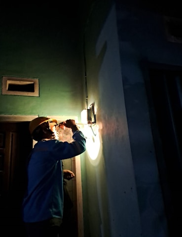 A person wearing a hard hat is inspecting a light fixture on a wall using a flashlight. The setting is dimly lit, suggesting an indoor environment, possibly under maintenance or construction. Shadows cast by the flashlight create a dramatic effect.