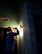 A person wearing a hard hat is inspecting a light fixture on a wall using a flashlight. The setting is dimly lit, suggesting an indoor environment, possibly under maintenance or construction. Shadows cast by the flashlight create a dramatic effect.