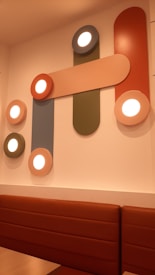 Geometric light fixtures are mounted on a wall with various shapes and colors. Circular lights are embedded in rounded rectangular panels of orange, peach, green, and blue. Below the installation is a cushioned brown bench, suggesting a seating area.