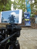 A smartphone mounted on a tripod showing the reflection of a palm tree and clouds on its screen. The background includes a banner with the text 'Let's up Hijrah' situated near a fence and surrounded by green leaves and trees.