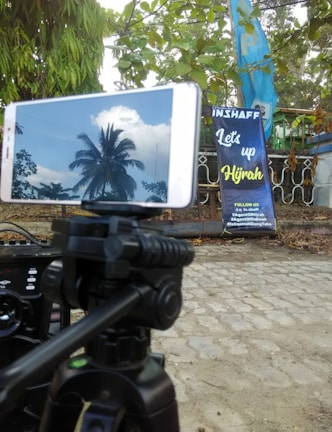A smartphone mounted on a tripod showing the reflection of a palm tree and clouds on its screen. The background includes a banner with the text 'Let's up Hijrah' situated near a fence and surrounded by green leaves and trees.