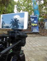 A smartphone mounted on a tripod showing the reflection of a palm tree and clouds on its screen. The background includes a banner with the text 'Let's up Hijrah' situated near a fence and surrounded by green leaves and trees.