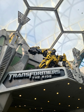 An entrance to an amusement park attraction featuring a large robot figure above a sign that reads 'Transformers The Ride.' The robot is yellow and black, designed to resemble a character from the Transformers franchise. The structure has a futuristic design with metallic elements and angular lines. The ceiling is made of glass panels arranged in a geometric pattern allowing natural light to illuminate the area.