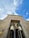 Two large statues resembling Anubis, the ancient Egyptian god with a jackal head, stand guard at the entrance of a structure with Egyptian-style architecture. The building facade is decorated with engravings and carvings, featuring traditional Egyptian motifs. Above, the sky is bright with scattered clouds.