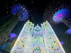 A vibrant and colorful display of lights forming intricate floral and geometric patterns, set under a dark night sky. Tall, tree-like structures with illuminated canopies add to the futuristic aesthetic.