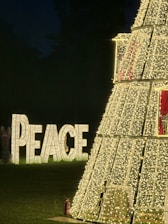 A large structure covered in numerous small white lights stands prominently in a dark outdoor setting. To the left of this structure, the word 'PEACE' is displayed in illuminated letters on the grass. The scene emanates a festive holiday atmosphere, possibly Christmas, indicated by the decorative lights and the peaceful message.