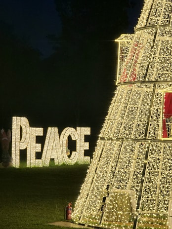 A large structure covered in numerous small white lights stands prominently in a dark outdoor setting. To the left of this structure, the word 'PEACE' is displayed in illuminated letters on the grass. The scene emanates a festive holiday atmosphere, possibly Christmas, indicated by the decorative lights and the peaceful message.