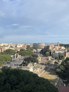 A panoramic view of a historic urban landscape featuring ancient ruins and modern architecture. The Colosseum is prominently visible amidst the surrounding buildings and lush greenery. A clear, blue sky dominates the background, and many people can be seen walking along the streets.