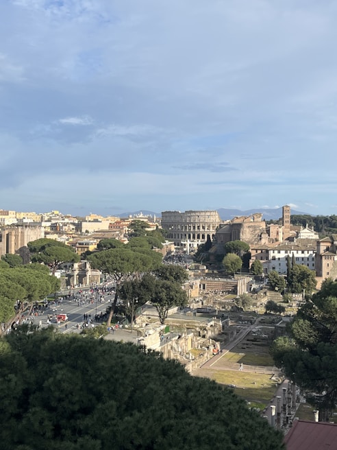 A panoramic view of a historic urban landscape featuring ancient ruins and modern architecture. The Colosseum is prominently visible amidst the surrounding buildings and lush greenery. A clear, blue sky dominates the background, and many people can be seen walking along the streets.