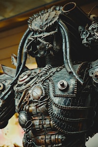 A detailed metallic sculpture resembling a futuristic or robotic warrior. The sculpture features intricate designs with various mechanical components, chains, and tube-like protrusions. Its overall appearance is rugged and complex, reflecting a high level of craftsmanship.