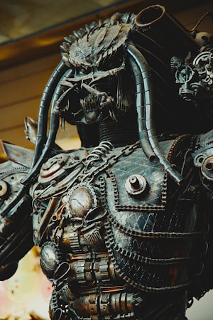 A detailed metallic sculpture resembling a futuristic or robotic warrior. The sculpture features intricate designs with various mechanical components, chains, and tube-like protrusions. Its overall appearance is rugged and complex, reflecting a high level of craftsmanship.