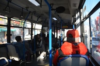 Inside a city bus, several passengers are seated, some are looking out the window while others are engaged in conversation. The interior features blue and gray seating and is illuminated by natural light coming through the large windows. A person in a bright red jacket and cap is prominently visible at the front.