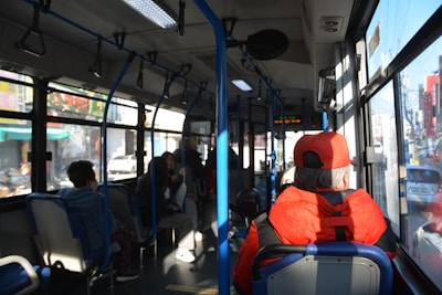 Inside a city bus, several passengers are seated, some are looking out the window while others are engaged in conversation. The interior features blue and gray seating and is illuminated by natural light coming through the large windows. A person in a bright red jacket and cap is prominently visible at the front.