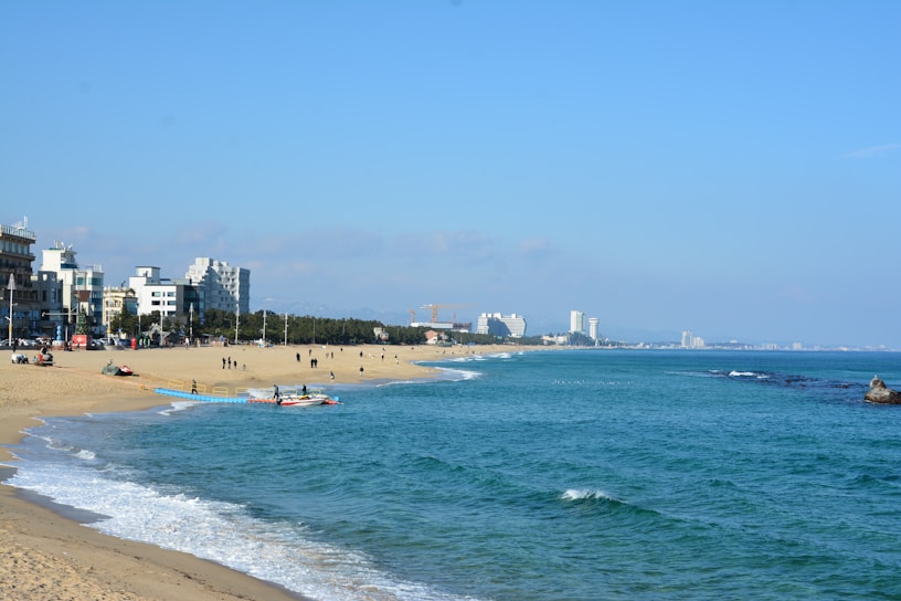 A sandy beach stretches alongside a vibrant blue ocean, with gentle waves lapping at the shore. People are scattered along the beach, some walking while others are near colorful boats. In the background, modern buildings and hotels line the coast, bordered by a lush green tree line.