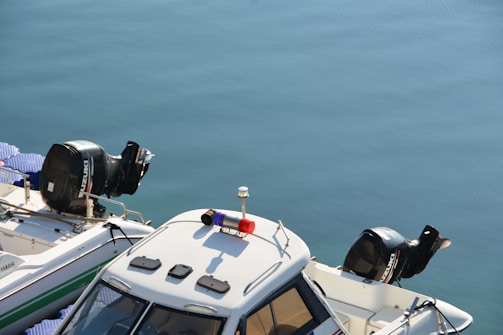 Two motorboats are docked side by side on calm blue water. The boats have visible outboard engines, one from Suzuki and the other from Yamaha. A light bar is mounted on the roof of one boat.