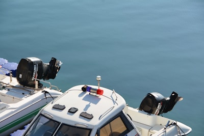 Two motorboats are docked side by side on calm blue water. The boats have visible outboard engines, one from Suzuki and the other from Yamaha. A light bar is mounted on the roof of one boat.