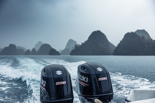 Twin Suzuki 200 outboard motors on the back of a boat with water spraying and a backdrop of rocky islands under an overcast sky. The scene is rainy, with visible water droplets.