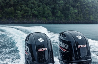 Two dark-colored outboard motors with the brand logo and number 200 are seen at the back of a boat traveling on water, surrounded by lush green forests in the background under rainy weather.