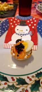 A small, round appetizer is placed on a decorative plate with a Christmas theme. The dish includes sliced carrots, a half-boiled egg, and a topping resembling caviar on a cream-covered base. In the background, a festive napkin with a snowman design adds to the holiday atmosphere.