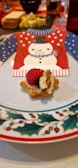 A festive holiday-themed plate with a decorative napkin featuring a snowman wearing a red and blue sweater pattern. In the foreground, a star-shaped appetizer is topped with a raspberry, a walnut, and a berry spread on a piece of toasted bread. There are blurred background elements, including a tray with sliced bread, cheese, and various condiments.