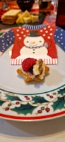 A festive holiday-themed plate with a decorative napkin featuring a snowman wearing a red and blue sweater pattern. In the foreground, a star-shaped appetizer is topped with a raspberry, a walnut, and a berry spread on a piece of toasted bread. There are blurred background elements, including a tray with sliced bread, cheese, and various condiments.