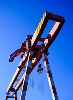 Several large industrial cranes with long arms and cables stand against a clear blue sky. The cranes are primarily painted in red and white with some yellow markings.