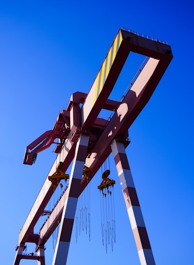 Several large industrial cranes with long arms and cables stand against a clear blue sky. The cranes are primarily painted in red and white with some yellow markings.